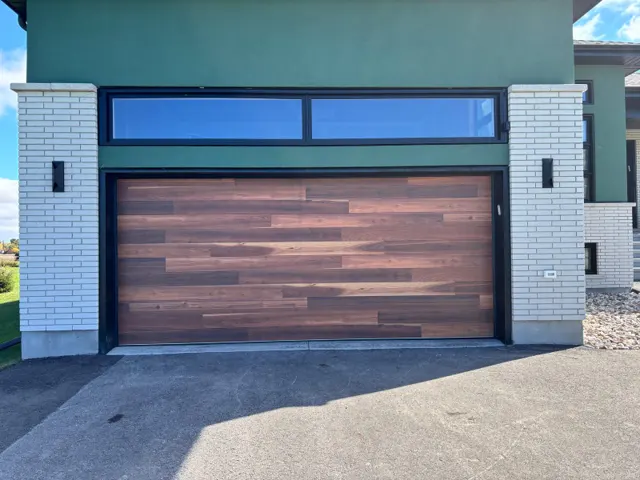 Double-width flush-panel garage door with a dark woodgrain finish, set between white brick pillars under a transom window on a modern home