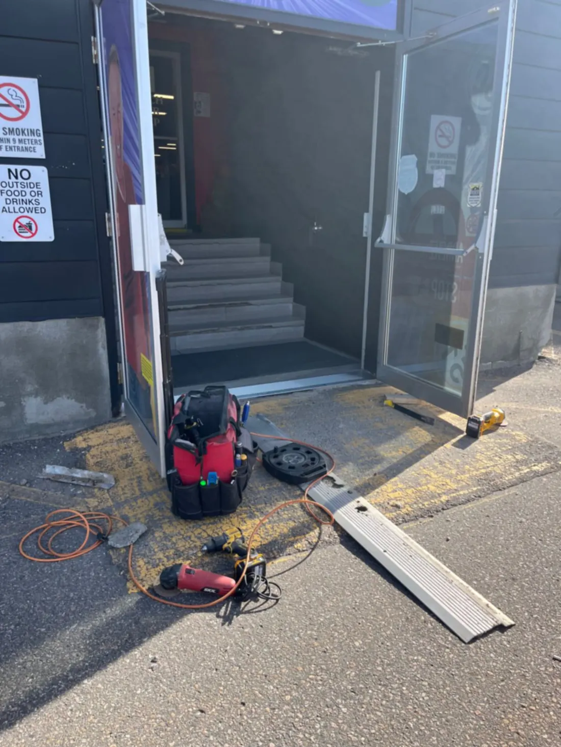 Glass storefront doors propped open during a threshold replacement, with a tool bag, grinder and the old threshold on the walkway
